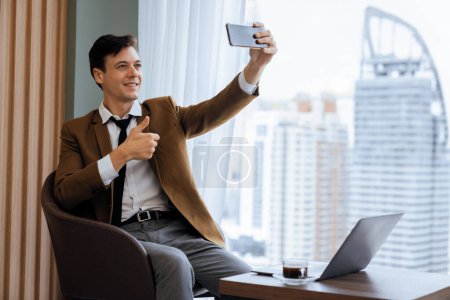 Photo for Professional businessman taking selfie near window with skyscraper while giving thumb up to camera. Skilled manager taking a photo while sitting at table with coffee cup and laptop. Ornamented. - Royalty Free Image