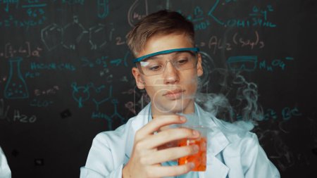 Photo for Closeup of boy inspect chemical solution while holding beaker at blackboard with chemical theory. Smart scientist doing experiment while analysis colored solution while wearing lab coat. Edification. - Royalty Free Image