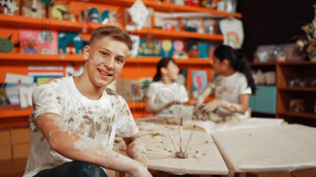 Photo for Highschool boy looking at camera while multicultural children modeling clay. Diverse student having pottery class together. Child smile while wearing white shirt with mud stained cloth. Edification. - Royalty Free Image