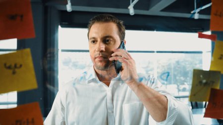 Photo for Professional businessman making phone call to customers while looking at marketing strategy idea written on colorful sticky notes. Project manager talking on phone analysis data on wall. Manipulator. - Royalty Free Image