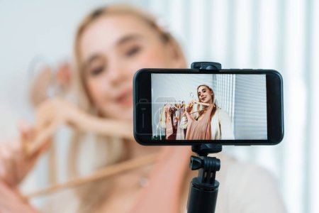 Téléchargez les photos : Jeune créateur de contenu social médial femme faire de la vidéo de mode. Le blogueur sourit et utilise le selfie du bâton de téléphone tout en faisant des vêtements en ligne persuasifs vendent vlog au public ou à l'abonné. Blithe - en image libre de droit
