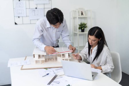 Photo for Professional male asian architect using ruler to measure house model length while young beautiful caucasian colleague using laptop to analyzed data on meeting table with house model. Immaculate. - Royalty Free Image