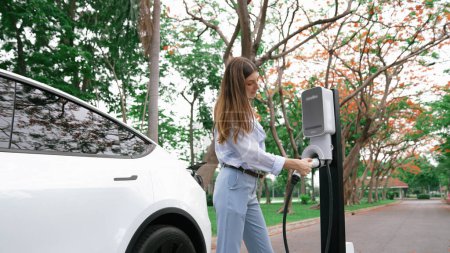 Woman recharging battery for electric car during road trip travel EV car in autumanl season natural forest or national park. Eco friendly travel during vacation and holiday. Exalt