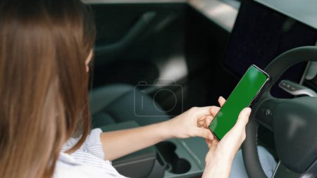 Photo for Holiday vacation road trip with environmental-friendly car concept. Eco-conscious woman on driver seat holding blank copyspace green screen smartphone for EV battery status. Exalt - Royalty Free Image