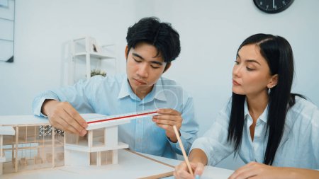 Professional male engineer measure house model by using ruler while beautiful cooperative coworker working together by writing on blueprint. Creative design and teamwork. Closeup. Immaculate.
