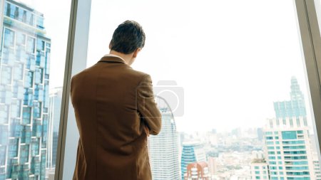 Téléchargez les photos : Arrière vue ambitieux homme d'affaires debout dans le bureau décoré regardant par la fenêtre sur l'horizon du paysage urbain. La détermination et l'ambition d'affaires poussent la carrière d'affaires vers un avenir brillant - en image libre de droit