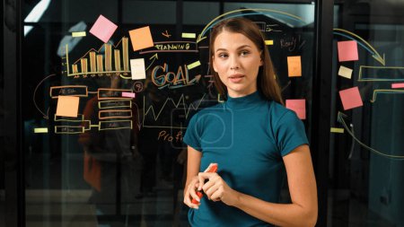 Photo for Skilled manager standing at glass wall while looking at camera. Professional caucasian businesswoman or female leader present marketing strategy while using sticky notes and mind mapping. Tracery - Royalty Free Image