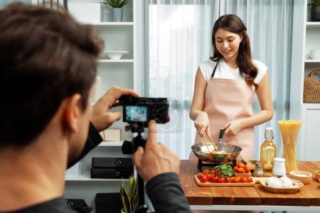 Photo for Cameraman recording to woman in chef influencer host cooking spaghetti with meat topped tomato sauce surrounded ingredients recipe, presenting special dish healthy food at modern studio. Postulate. - Royalty Free Image