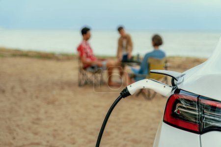 Téléchargez les photos : Voyage alternatif de vacances en famille voyageant par la plage avec batterie de recharge de voiture électrique de la station de charge EV avec la famille floue profitant du fond marin. Perpétuel - en image libre de droit