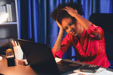Téléchargez les photos : Femme d'affaires africaine, portant une chemise rose ayant mal à la tête avec migraine, assise à l'ordinateur portable avec position penchée. Concept de forme de travail à la maison avec pression et tension. Tastemaker. - en image libre de droit