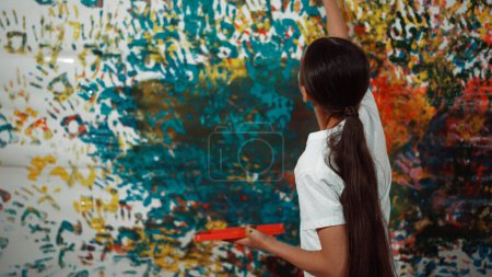 Photo for Young happy girl holding the color tray and paint colorful stained wall to express idea and enhance imagination with hand print. Caucasian girl stamp hand to decorate room in art lesson. Edification. - Royalty Free Image