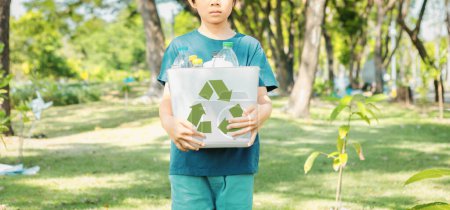 Fröhlicher junger asiatischer Junge, der die Mülltonne mit dem Recycling-Symbol auf dem Tageslicht-Naturpark hält und das Recycling, die Reduzierung und Wiederverwendung von Abfällen fördert, um zukünftigen Generationen ein ökologisch nachhaltiges Bewusstsein zu vermitteln. Kreisel