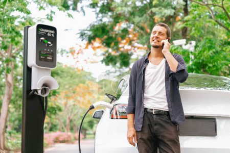Photo for Man talking on smartphone while recharging electric car battery charging from EV charging station during vacation holiday road trip at national park or autumnal forest. Exalt - Royalty Free Image