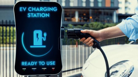 Photo for Businessman pull and hold EV charger plug form electric car charging station at city car park area background. Futuristic clean sustainable energy and EV car technological advancement.Peruse - Royalty Free Image