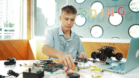 Highschool-Junge repariert Auto-Modell mit Laptop und Elektrowerkzeug auf dem Tisch platziert. Jugendliche inspizieren in Großaufnahme den Roboterbau, während sie elektronische Geräte im MINT-Unterricht einsetzen. Erbauung.