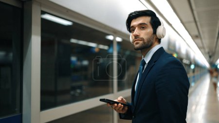 Photo for Project manager wearing headphone at train station while holding mobile phone for choosing song. Smart business man listening relaxing music while waiting for train with blurred background. Exultant. - Royalty Free Image