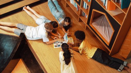 Photo for Group of diverse children lying down in circle while reading a book at library. Top view of girl sharing a magazine while pointing at interested topic and talking with lovely friends. Edification. - Royalty Free Image