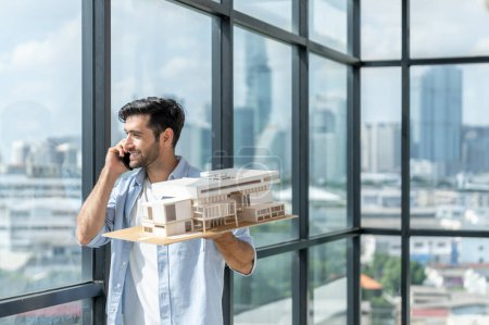 Photo for Handsome skilled architect engineer holds architectural model while inspect house model. Professional interior designer checking house construction while standing near window with city view. Tracery. - Royalty Free Image