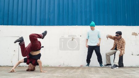 Young street dancer dancing in hip hop style with multicultural friends cheering behind at wall. Attractive handsome man stretch arms at street with blue background. Outdoor sport 2024. Endeavor.