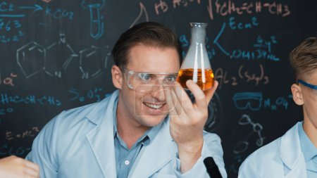 Kluger Lehrer hält Lösung im Becher, während Schüler im naturwissenschaftlichen MINT-Unterricht Experimente an der Tafel mit chemischer Theorie machen. Fachkundige Wissenschaftler untersuchen chemische Flüssigkeiten im Labor. Erbauung.