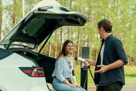 Photo for Lovely young couple recharging battery for electric car while relaxing and sitting on the trunk during road trip travel EV car in autumnal forest. Eco friendly travel on vacation during autumn. Exalt - Royalty Free Image