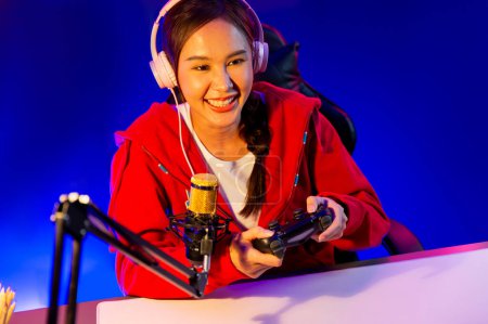 Téléchargez les photos : Canal hôte de sourire belle fille asiatique streamer avec joystick jouer à un jeu en ligne casque coller parler avec les téléspectateurs médias en ligne. Joueurs d'équipe qualifiés Esport dans la salle néon. Stratagème. - en image libre de droit