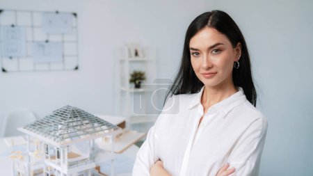 Photo for Close up portrait of young beautiful professional architect engineer cross arms and poses confidently with house model at white modern office with blueprint. Creative living and design. Immaculate. - Royalty Free Image