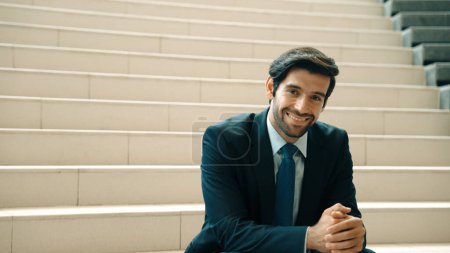 Téléchargez les photos : Homme d'affaires habile souriant regardant la caméra tout en étant assis sur les escaliers. Jeune chef de projet professionnel sourire à la caméra tout en tenant le téléphone mobile à l'extérieur avec fond flou. Exultant. - en image libre de droit