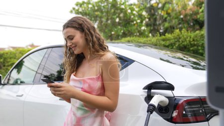 Photo for Young woman use smartphone to pay for electricity at public EV car charging station in nature. Modern environmental and sustainable automobile transportation lifestyle with EV vehicle. Synchronos - Royalty Free Image