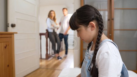 Gestresstes und unglückliches junges Mädchen schmiegt sich in die Ecke, verdeckt ihre Ohren, während ihre Eltern im Hintergrund streiten. Häusliche Gewalt zu Hause und traumatische Kindheit entwickeln sich zu Depressionen. Synchronos