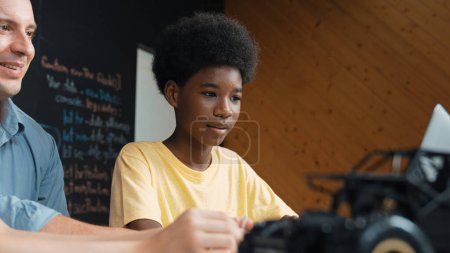 Photo for African boy using laptop to program code or prompt with car model placed while teacher and caucasian friend study about robotic construction. Student study about car model structure. Edification. - Royalty Free Image