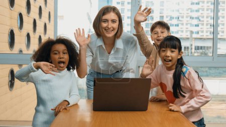 Photo for Happy caucasian teacher and multiethnic children waving hands to camera in casual uniform. Smart mentor and diverse student looking at laptop while greeting by moving hands or say good bye. Erudition. - Royalty Free Image