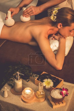 Téléchargez les photos : Chaud à base de plantes balle spa massage du corps, masseur compresse doucement sac à herbes sur le corps de la femme. Tranquille et sérénité des loisirs aromathérapie dans l'éclairage chaud des bougies au salon de spa. Tranquillité - en image libre de droit