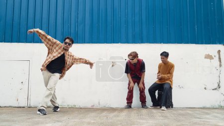 Young street dancer dancing in hip hop style with multicultural friends cheering behind at wall. Attractive handsome man stretch arms at street with blue background. Outdoor sport 2024. Endeavor.