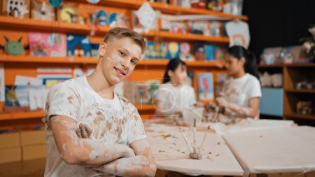 Photo for Smiling boy looking at camera and crossing arm with confident at workshop while diverse student having pottery class together. Happy caucasian student smile while pose with arm folded. Edification. - Royalty Free Image