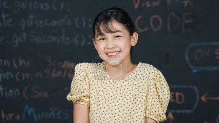 Photo for Cute happy girl smiling to camera while standing at blackboard with engineering prompt or coding, programing system written in STEM technology classroom. Smart student looking at camera. Edification. - Royalty Free Image