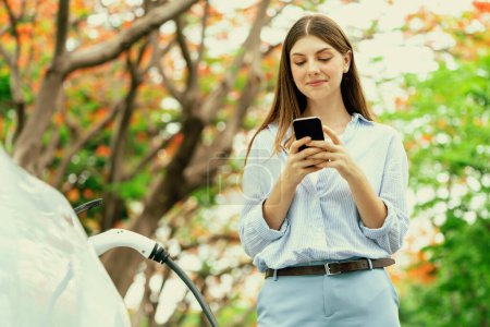 Jeune femme utilisant smartphone application bancaire en ligne pour payer la charge de la batterie de voiture électrique de la station de charge EV pendant les vacances d'automne voyage dans le parc national ou la forêt automnale. Exalt.
