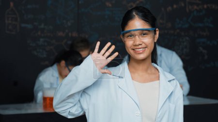 Photo for Cute girl waving hands to camera while people doing experiment at laboratory. Young cute student standing blackboard with chemical theory with blurring background at STEM science class. Edification. - Royalty Free Image