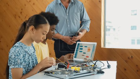 Photo for Teacher standing while explain coding and robotic construction. Diverse teenager fixing mother board and using electronic tool while looking at laptop display programing code screen. Edification. - Royalty Free Image