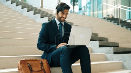 Téléchargez les photos : Homme d'affaires professionnel assis à l'escalier tout en travaillant sur un ordinateur portable. Gestionnaire de projet compétent écouter de la musique à partir du casque et vérifier les e-mails et la discussion sur le plan de marketing. En plein air. Exultant. - en image libre de droit