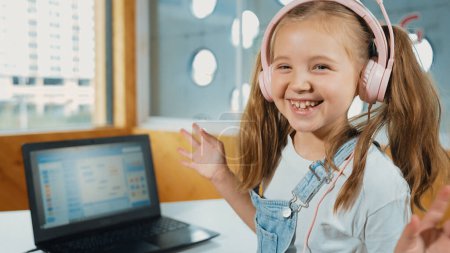 Chica sonriente mirando mientras agita la mano a la cámara con el ordenador portátil colocado en la mesa. Niño que usa auriculares sonriendo mientras la pantalla del portátil muestra el programa de programación o codificación del sistema en la clase STEM. Erudición.