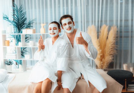 Téléchargez les photos : Couple heureux en peignoir avec masque crème visage profitant d'une ambiance sereine de spa salon resort ou hôtel pendant les vacances. Soin du visage choyante spa et soins de la peau avec relaxation essence. Tranquillité - en image libre de droit