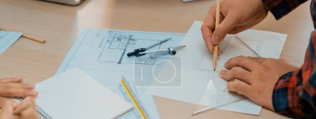 Téléchargez les photos : Architecte professionnel dessiner à la main un plan en utilisant règle mesurant la longueur sur la table avec l'équipement architectural et le plan disperser autour au bureau moderne. Gros plan. Délimitation. - en image libre de droit