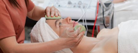 Schöne junge Frauen liegen auf einem Wellnessbett, während sie Gesichtsmassage von einem professionellen Arzt erhalten. Attraktive Frau mit schöner Haut, umgeben von elektrischer Gesichtsmaschine. Gelassenheit.