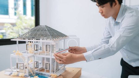 Téléchargez les photos : Image rapprochée d'un ingénieur architecte asiatique professionnel se concentrant sur la mesure modèle de maison moderne au bureau moderne blanc. Concentrez-vous. Concept créatif de vie et de design. Immaculée. - en image libre de droit