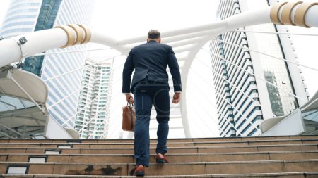 Téléchargez les photos : Homme d'affaires caucasien intelligent montant les escaliers entouré d'une vue urbaine. Vue arrière de gestionnaire de projet ambitieux trouver un emploi stimulant ou une opportunité d'investissement à la ville avec un bâtiment moderne. Urbane. - en image libre de droit