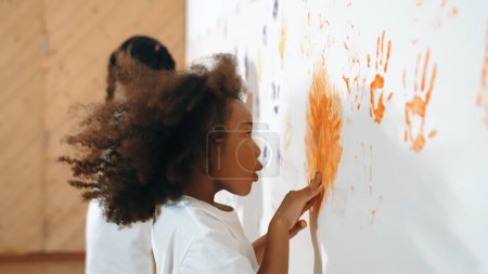 Téléchargez les photos : Africaine fille peindre le mur avec des mains colorées teintées avec un ami. Groupe d'apprenants multiculturels debout devant le mur tout en utilisant la main pour créer des œuvres d'art. Concept d'activité créative. Erudition. - en image libre de droit