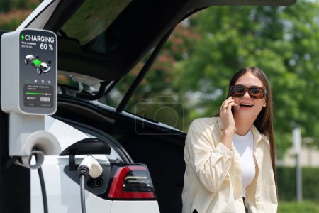Photo for Young woman recharge her EV electric vehicle at green city park parking lot while talking on phone. Sustainable urban lifestyle for environment friendly EV car with battery charging station. Expedient - Royalty Free Image
