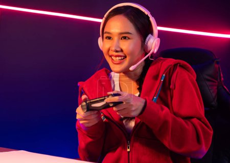 Photo for Host channel of smiling beautiful Asian girl streamer with joystick playing online game wearing headphones paste talking with viewers media online. Esport skilled team players in neon room. Stratagem. - Royalty Free Image