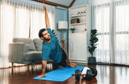 Foto de Hombre flexible y hábil en ropa deportiva haciendo posición de yoga en postura de meditación sobre colchoneta de ejercicio en casa. Gaiety sana estilo de vida casero del yoga con mente pacífica y serenidad. - Imagen libre de derechos
